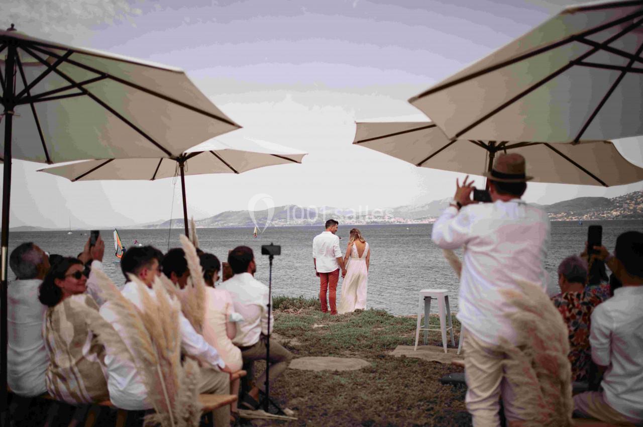 Un couple échange ses vœux face à la mer, entouré d'invités assis sous des parasols.
