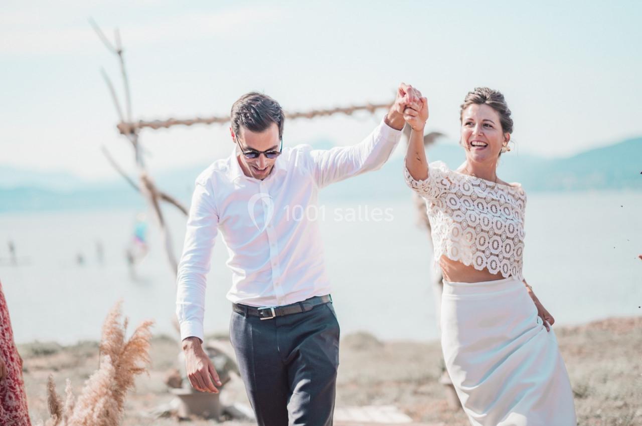 Un couple souriant marche main dans la main près d'une plage, sous un ciel clair.