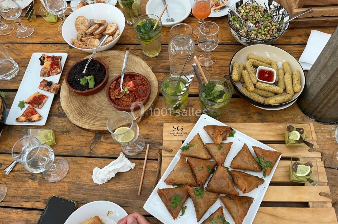 Table en bois garnie de plats variés : samoussas, mezzés, crudités, boissons et verres vides ou partiellement remplis.
