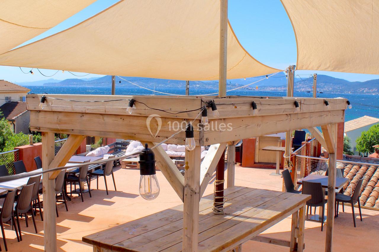 Table en bois sous des voiles d'ombrage beige sur une terrasse avec vue sur un paysage côtier en arrière-plan.