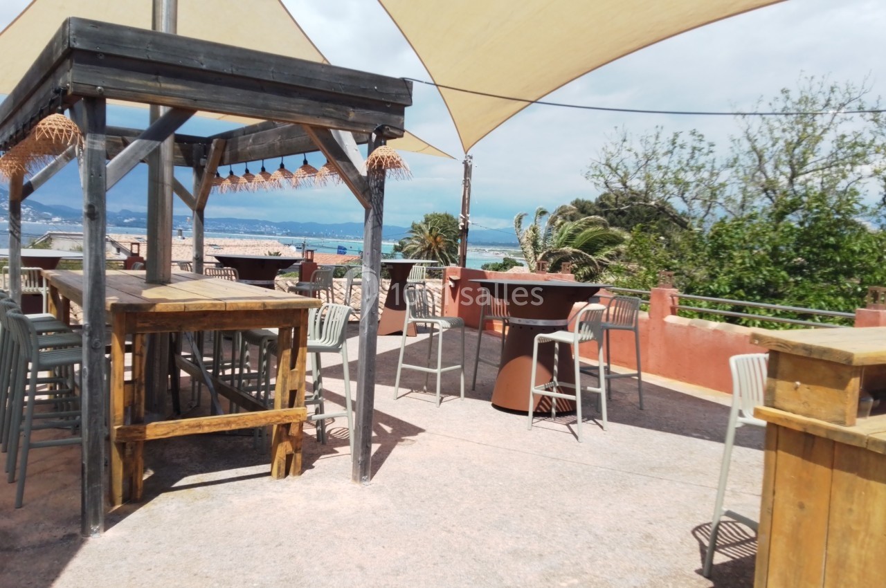 Terrasse extérieure avec tables en bois, chaises et parasols, offrant une vue sur la mer et des arbres en arrière-plan.