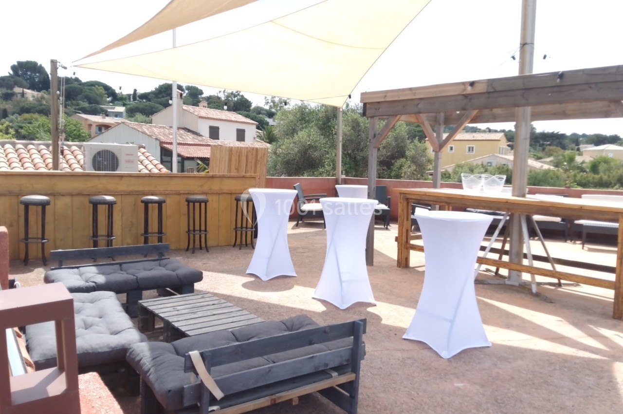 Terrasse aménagée avec canapés, tables hautes, tabourets et pergola, entourée de maisons et végétation.