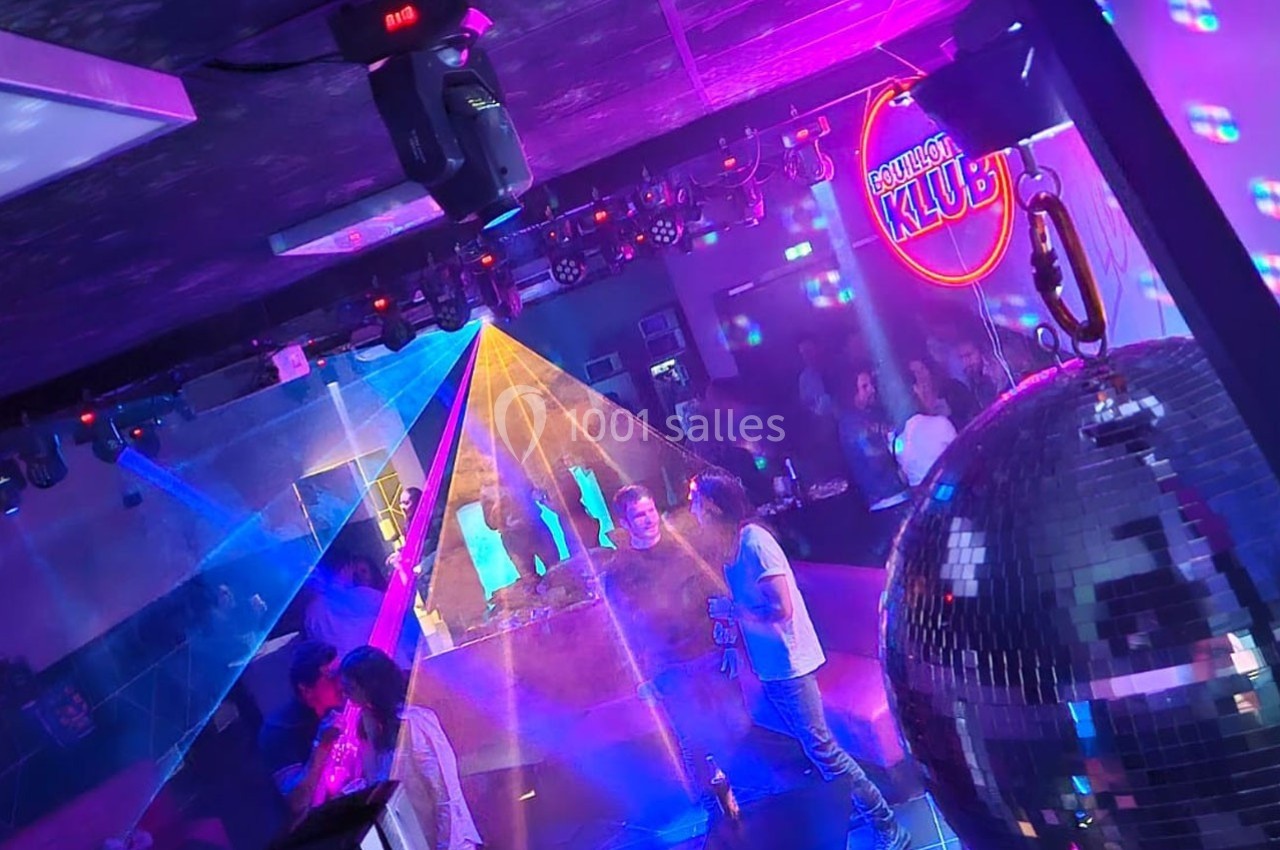 Des personnes dansent dans une boîte de nuit avec des jeux de lumière colorés, un miroir disco et un bar en arrière-plan.