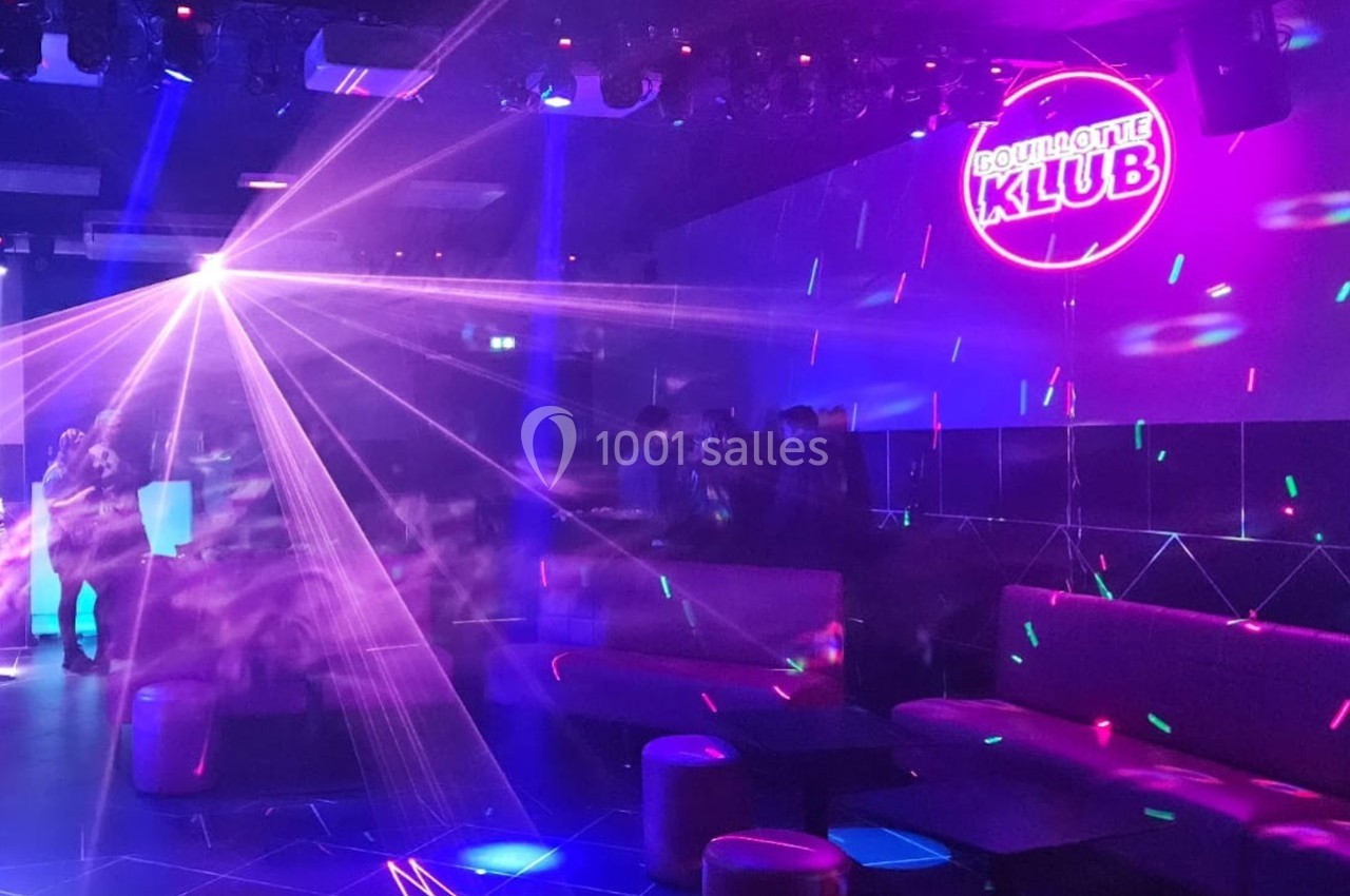 Salle de discothèque avec jeux de lumières colorées, néons et quelques personnes autour de tables basses.