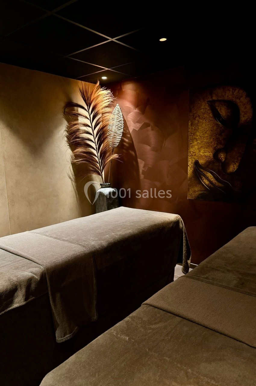 Salle de massage tamisée avec deux tables recouvertes de draps bruns et une décoration murale apaisante.