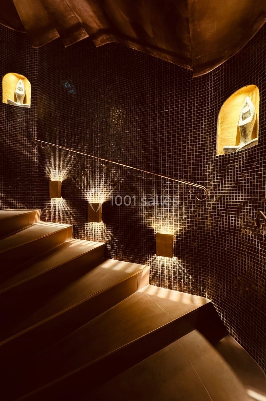 Escalier en colimaçon avec des murs recouverts de mosaïques sombres, éclairé par des niches lumineuses.