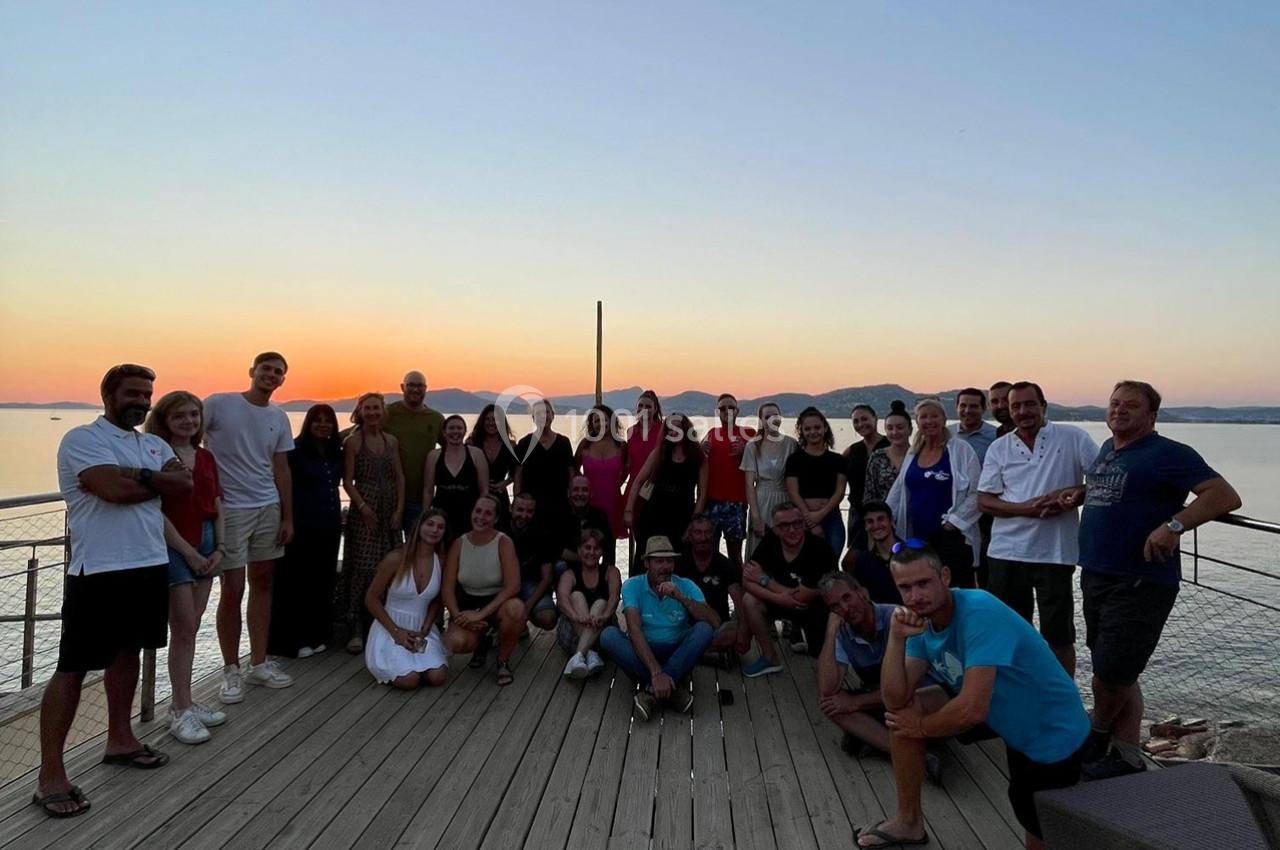 Groupe de personnes posant sur une terrasse en bois au bord de l'eau, avec un coucher de soleil en arrière-plan.