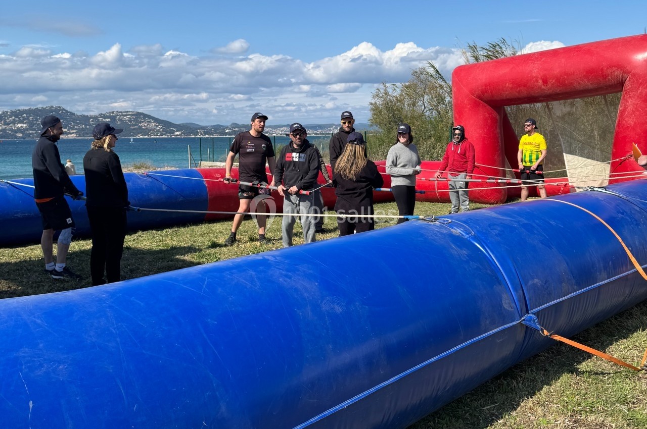 Des personnes debout autour d'un terrain gonflable bleu et rouge en plein air, près d'un plan d'eau.