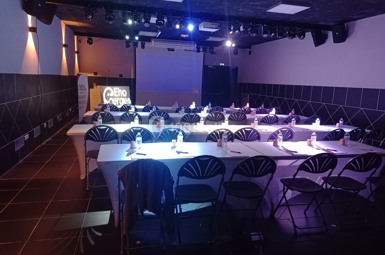 Salle de conférence sombre avec des tables alignées, chaises pliantes et projecteur affichant un logo sur un écran.