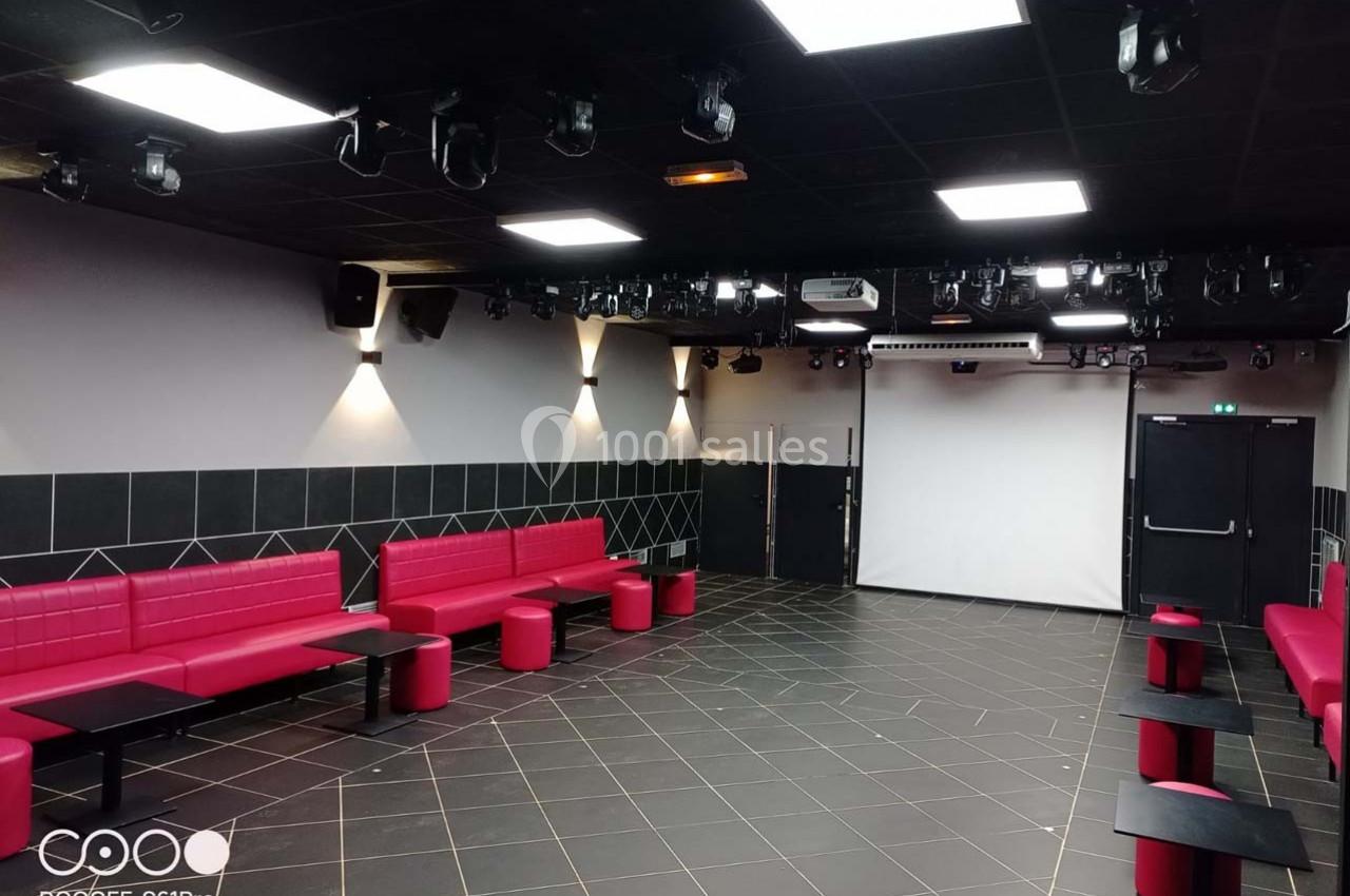 Salle moderne avec banquettes rouges, tables noires, éclairage au plafond et un écran blanc au fond.