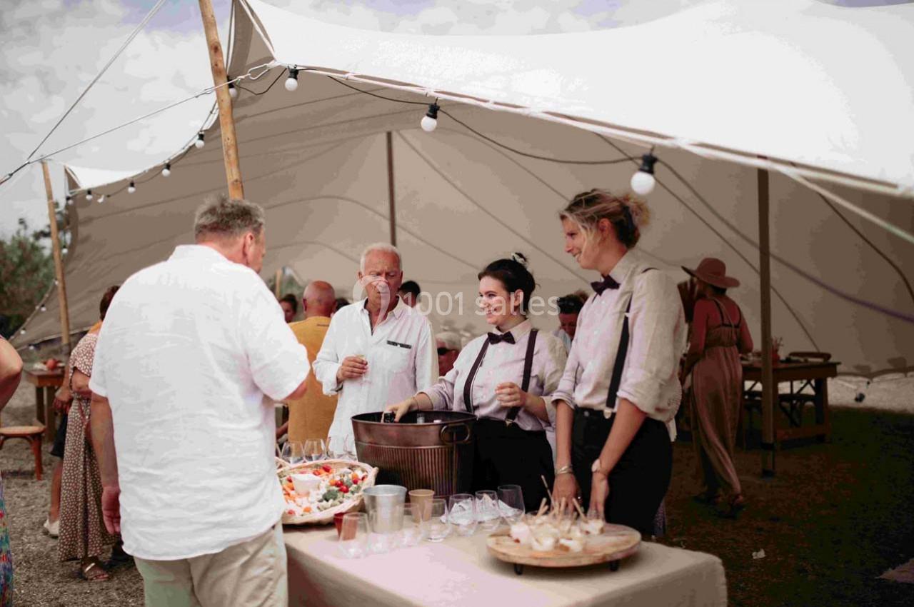 Des serveurs en tenue formelle servent des boissons à des invités sous une tente blanche lors d'un événement en extérieur.