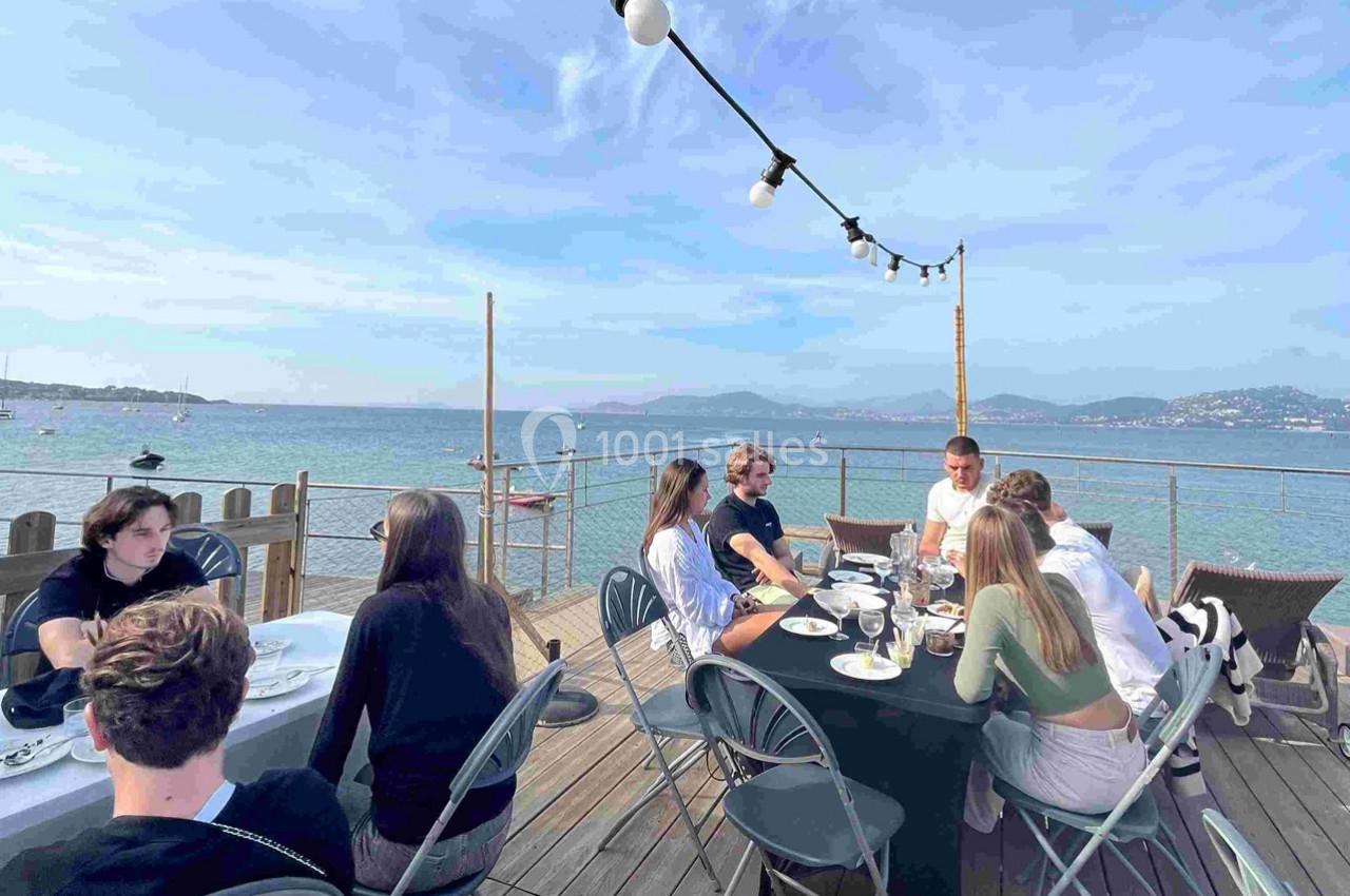 Des personnes dînent en terrasse en bord de mer, avec une vue sur l'eau et les montagnes à l'horizon.