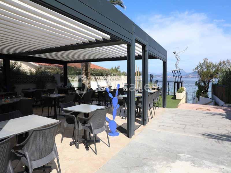 Terrasse couverte d'un restaurant avec des tables et chaises, vue sur la mer et sculpture moderne à proximité.