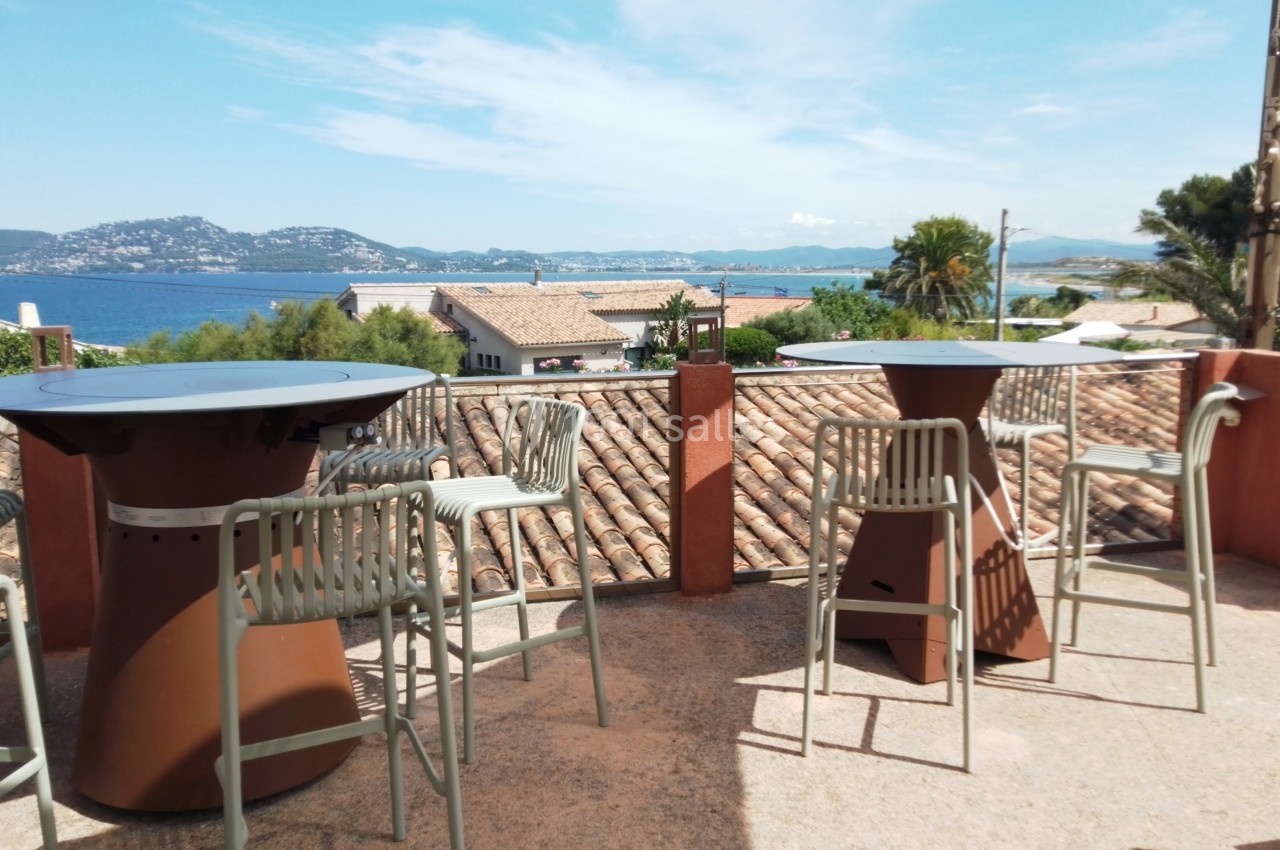 Terrasse avec tables hautes, chaises en métal et vue sur des toits, la mer et des collines sous un ciel dégagé.