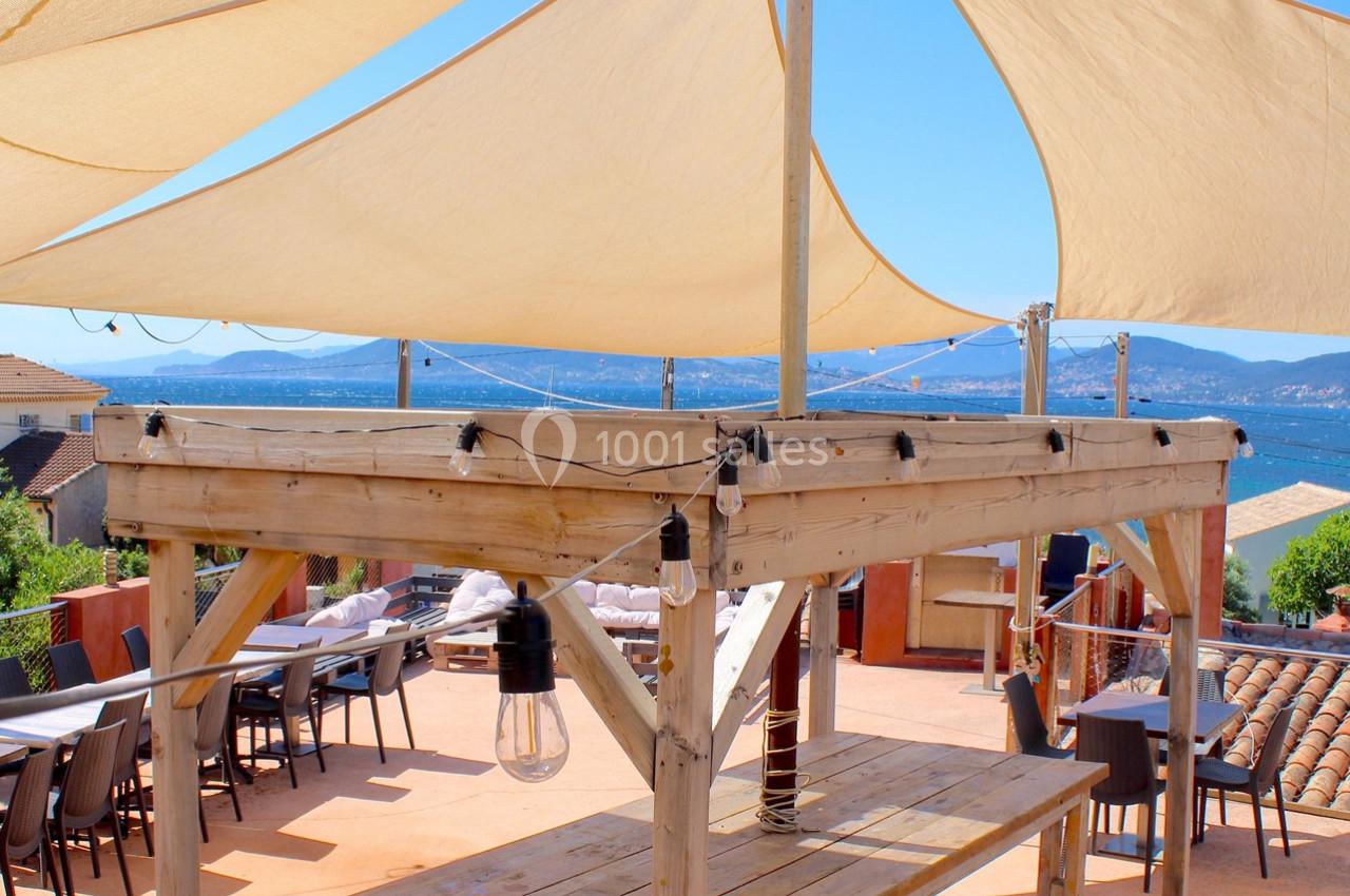 Terrasse ensoleillée avec voiles d'ombrage, vue sur la mer et les collines, tables et chaises disposées en extérieur.