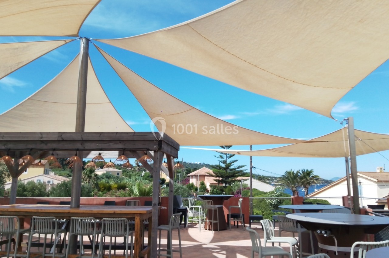 Terrasse ensoleillée avec voiles d'ombrage, tables et chaises, entourée de végétation et maisons en arrière-plan.