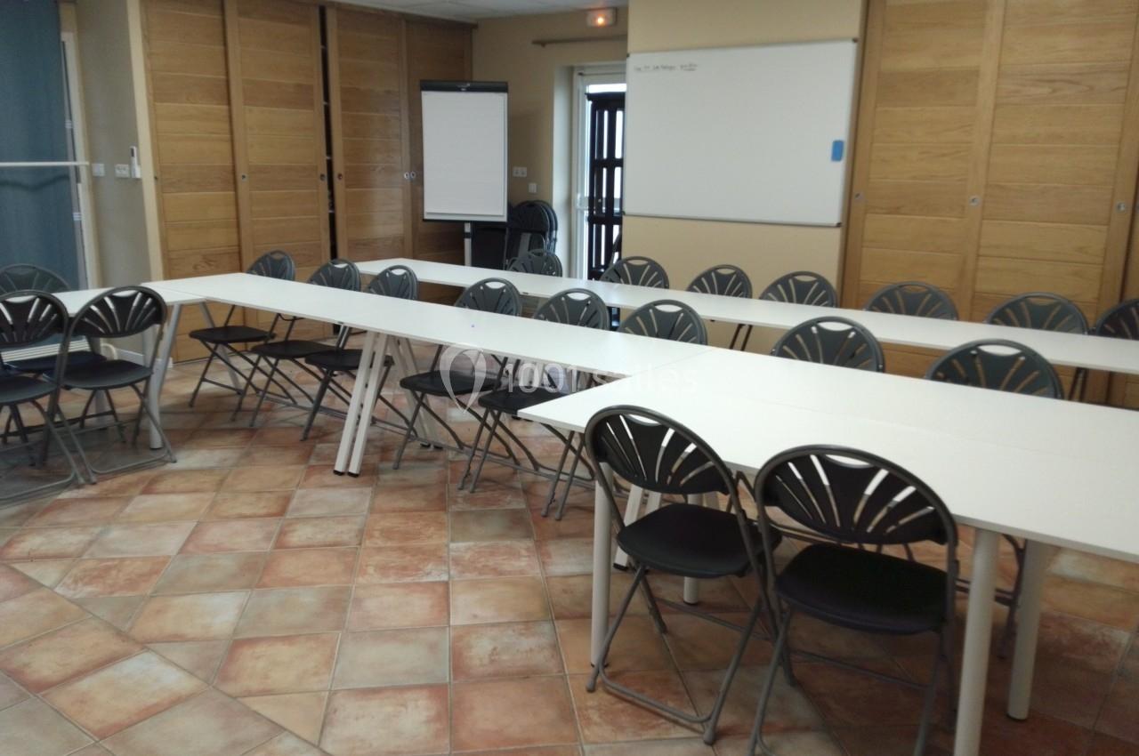 Salle de réunion avec des tables blanches disposées en U, des chaises noires et un tableau blanc au mur.