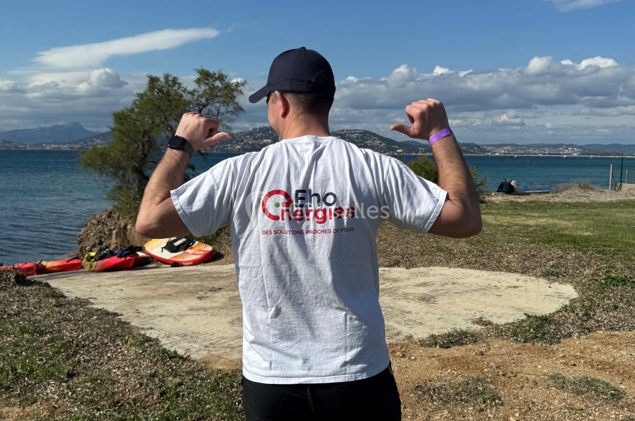 Un homme de dos portant un t-shirt blanc avec un logo, debout sur une pelouse près d'un lac par temps ensoleillé.