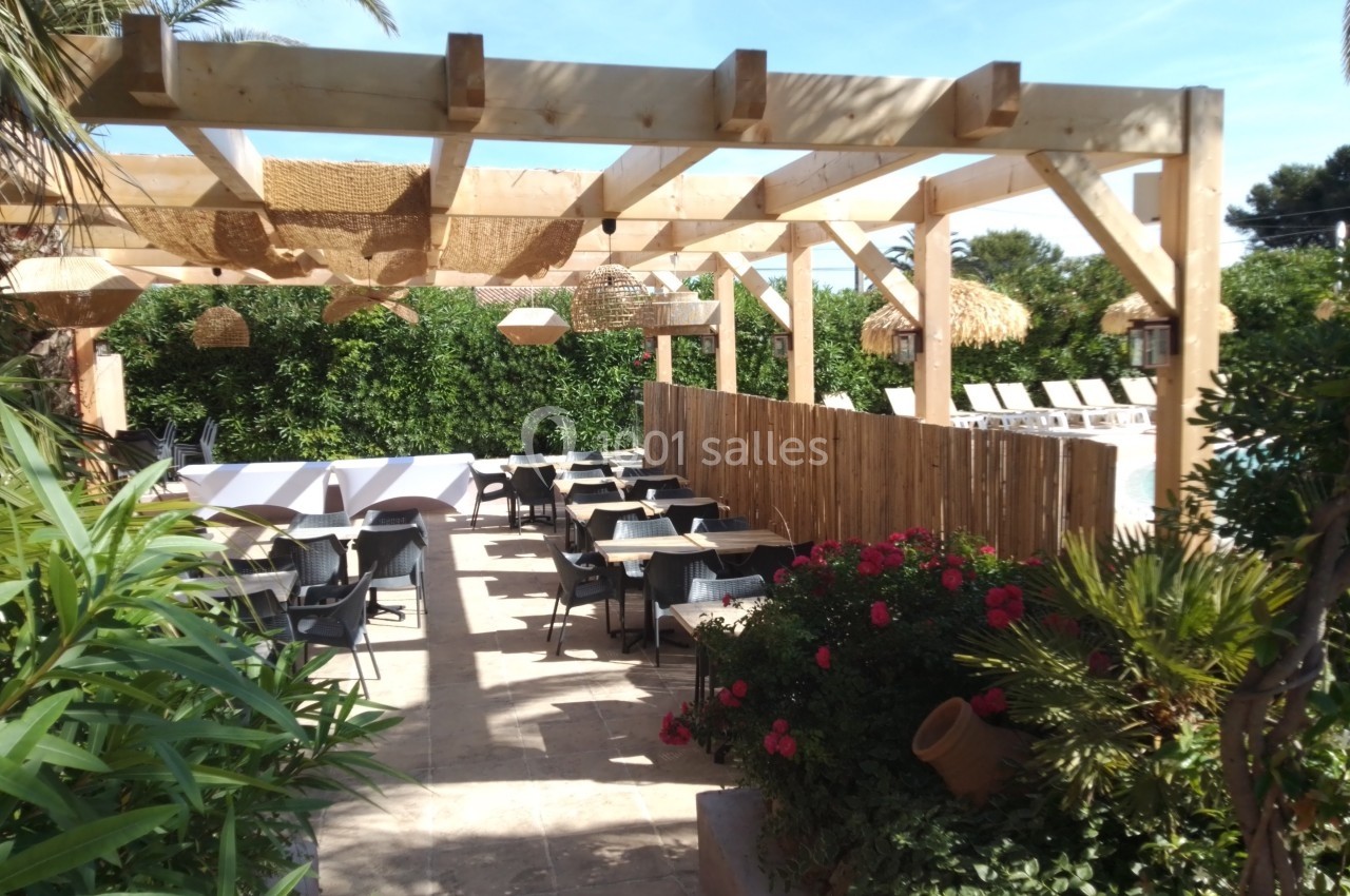 Terrasse extérieure avec pergola en bois, tables et chaises, entourée de végétation et fleurs colorées.