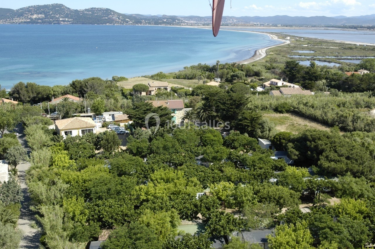 Vue aérienne d'un paysage côtier avec des maisons entourées de végétation dense, une plage et la mer en arrière-plan.
