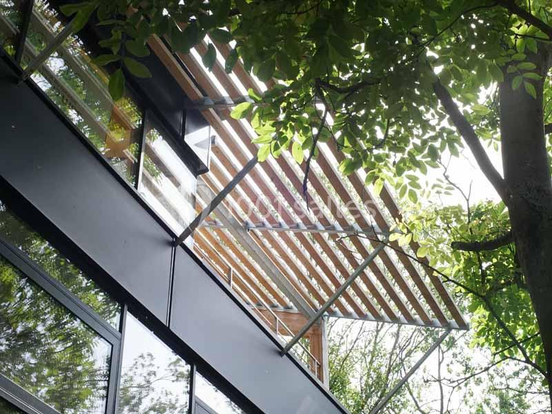 Façade d'un bâtiment moderne avec des lames en bois formant une pergola, entourée de végétation.