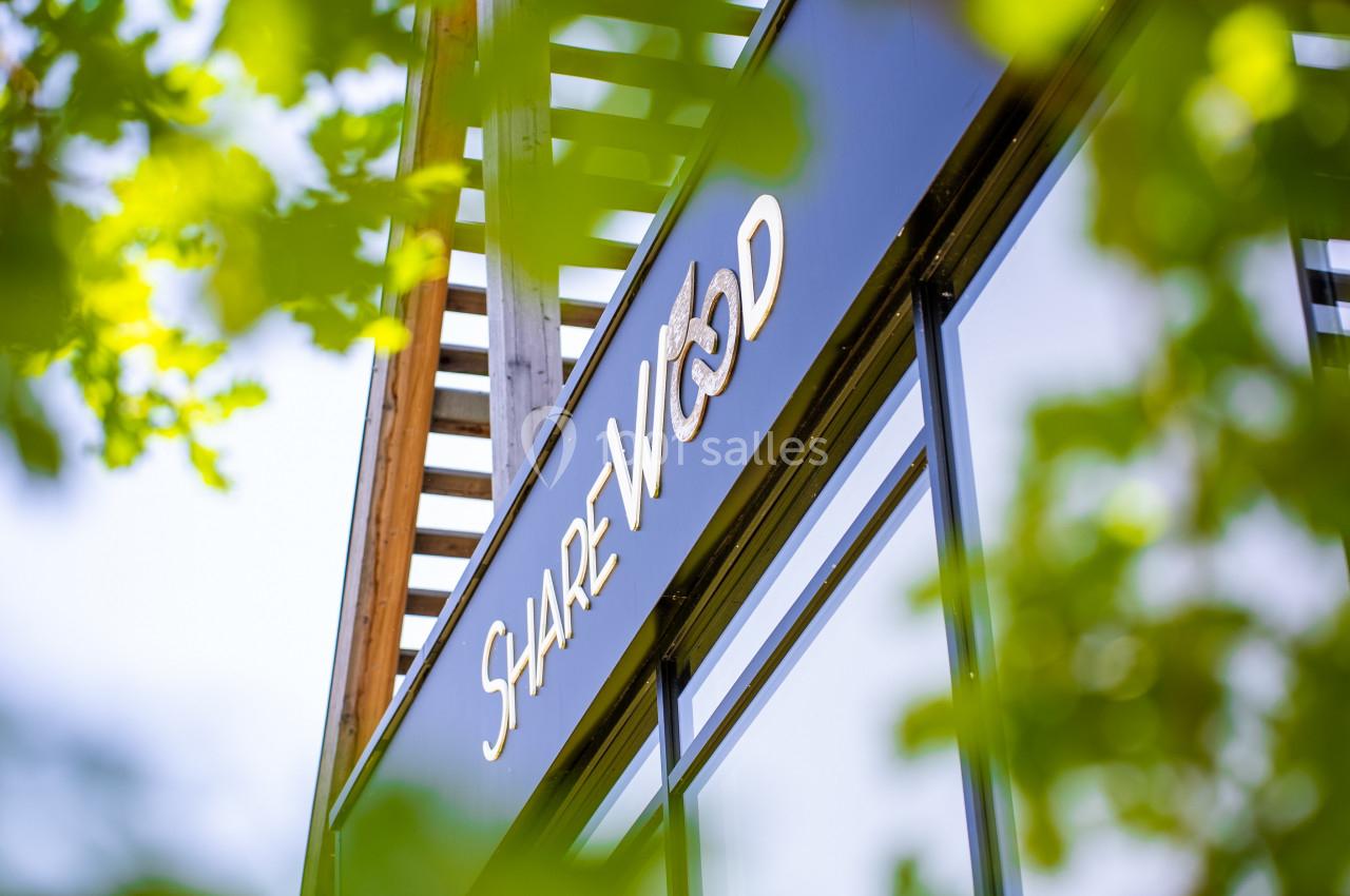 Façade d'un bâtiment moderne avec une enseigne ’Sharewood’, entourée de feuillage vert au premier plan.