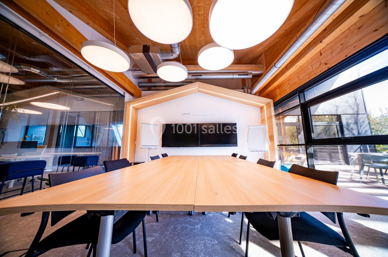 Salle de réunion moderne avec une grande table en bois, des chaises noires, des écrans et un éclairage suspendu.