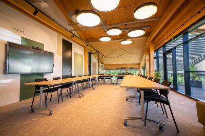 Salle de réunion lumineuse avec table en bois, quatre chaises noires, écran mural et cloison vitrée donnant sur l'extérieur.