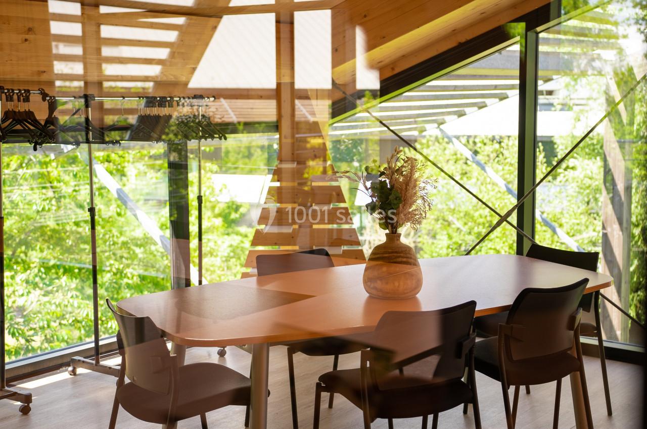 Salle lumineuse avec une grande table, des chaises, des murs vitrés et une vue sur un environnement verdoyant.