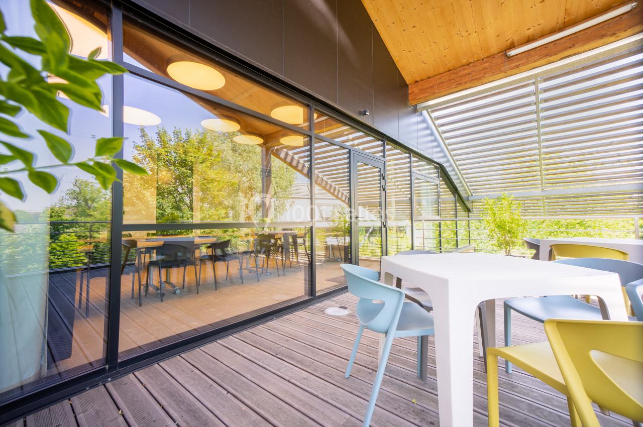 Terrasse en bois avec table et chaises colorées, donnant sur une salle vitrée éclairée et entourée de verdure.