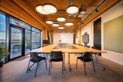 Salle de réunion lumineuse avec table en bois, quatre chaises noires, écran mural et cloison vitrée donnant sur l'extérieur.