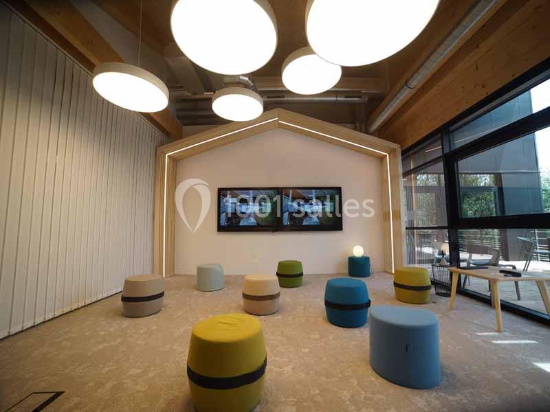 Salle moderne avec des poufs colorés, des écrans muraux, des luminaires suspendus et une grande baie vitrée.