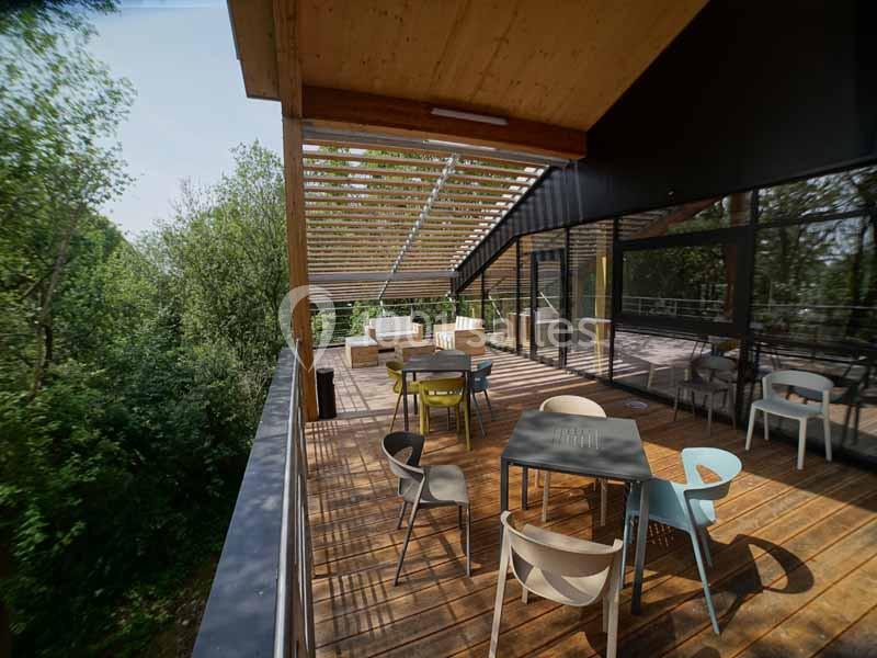 Terrasse en bois avec tables et chaises colorées, partiellement ombragée par une pergola, entourée de végétation.