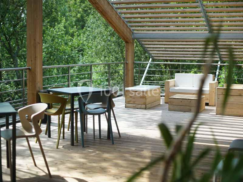 Terrasse en bois avec tables, chaises et banquettes, entourée de verdure et partiellement couverte par une pergola.
