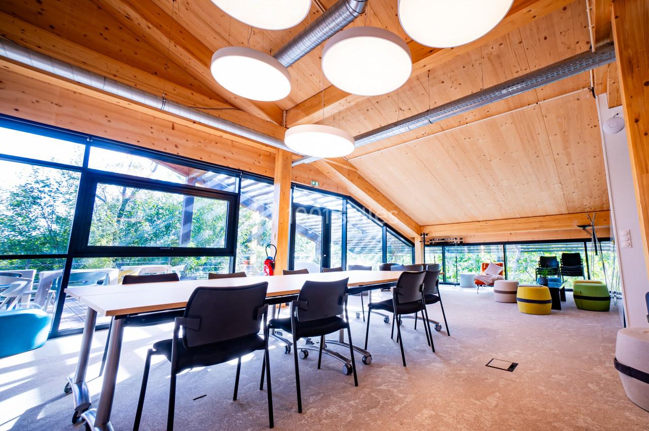 Salle de réunion lumineuse avec table, chaises noires, plafond en bois et grandes baies vitrées donnant sur un espace…