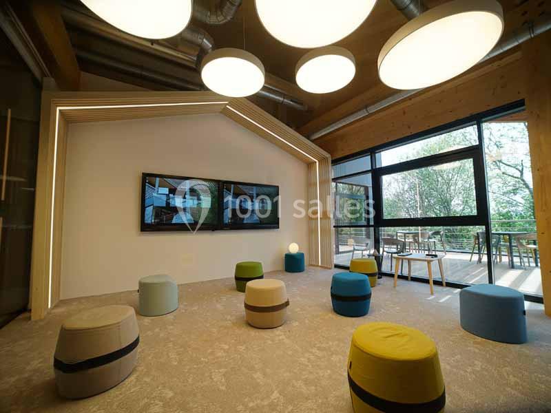 Espace intérieur moderne avec poufs colorés, écrans muraux et grandes fenêtres donnant sur une terrasse et des arbres.