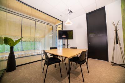 Salle de réunion lumineuse avec table en bois, quatre chaises noires, écran mural et cloison vitrée donnant sur l'extérieur.