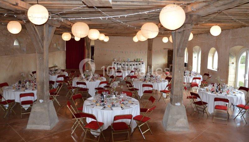 Salle de réception rustique avec tables rondes dressées, chaises rouges et lanternes suspendues au plafond en bois.