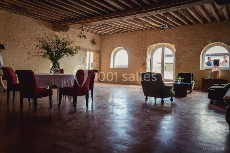 Salle lumineuse avec murs en pierre, poutres apparentes, mobilier éclectique et grandes fenêtres en arc.