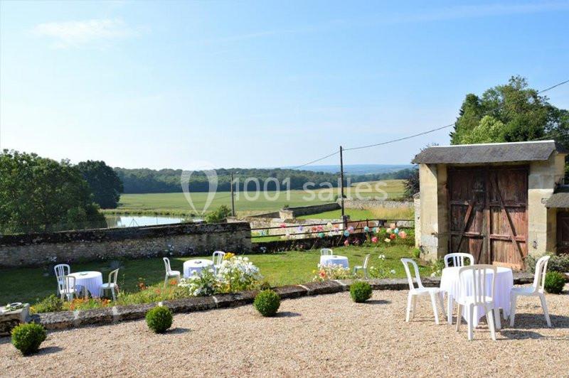 Jardin avec tables et chaises blanches, vue sur un paysage champêtre avec étang et arbres en arrière-plan.