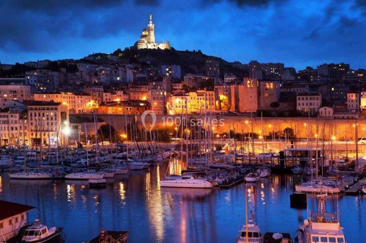 Vue nocturne du Vieux-Port de Marseille avec des bateaux amarrés et la basilique Notre-Dame de la Garde illuminée en arrière…