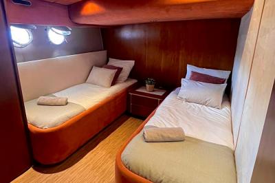 Cabine de bateau avec deux lits simples, linge de lit beige et rouge, éclairage intégré et hublots.