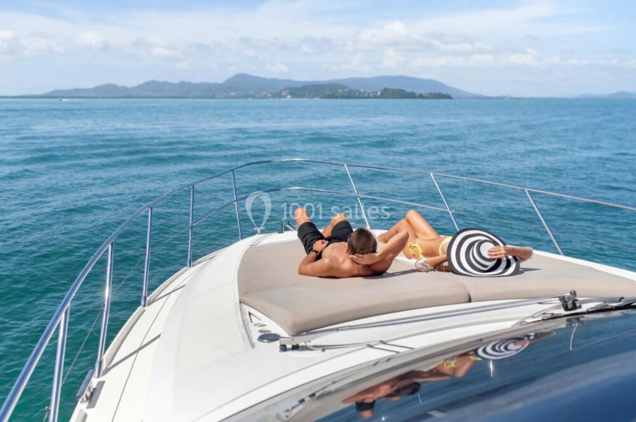 Un couple allongé sur le pont d'un yacht, profitant du soleil avec une vue sur la mer et des îles au loin.