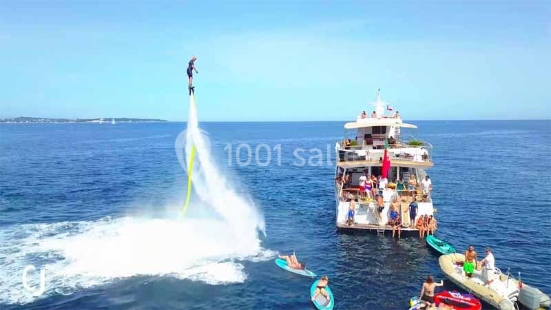 Un homme pratique le flyboard au-dessus de l'eau près d'un yacht entouré de personnes sur des paddleboards.