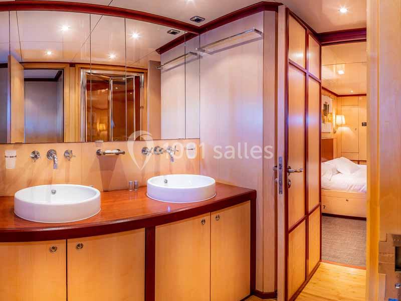 Salle de bain avec deux lavabos, grand miroir et vue partielle sur une chambre avec lit au fond.