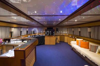 Un bateau à moteur tracte une bouée gonflable sur une mer calme, avec un yacht et une côte boisée en arrière-plan.