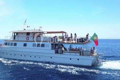 Un bateau à moteur tracte une bouée gonflable sur une mer calme, avec un yacht et une côte boisée en arrière-plan.