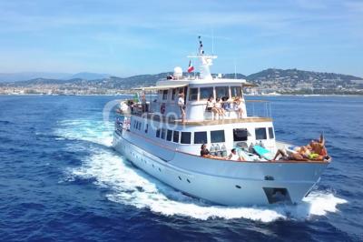 Un bateau à moteur tracte une bouée gonflable sur une mer calme, avec un yacht et une côte boisée en arrière-plan.