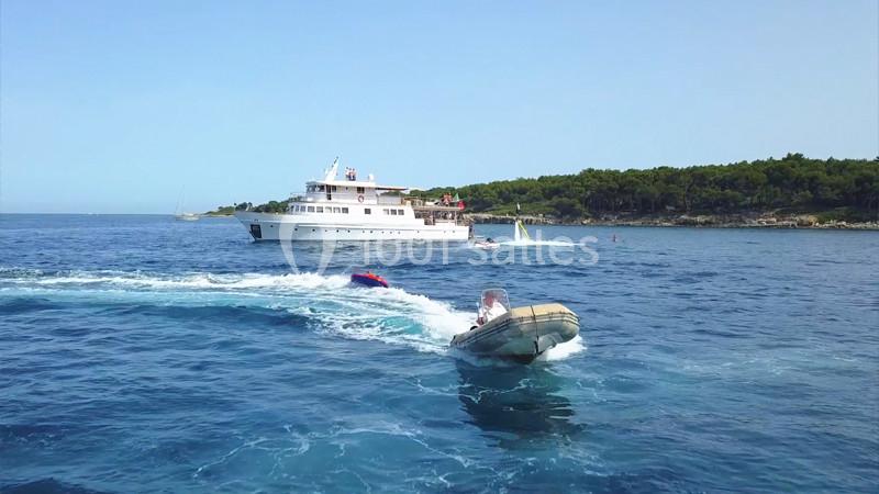 Un bateau à moteur tracte une bouée gonflable sur une mer calme, avec un yacht et une côte boisée en arrière-plan.