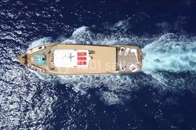 Un bateau à moteur tracte une bouée gonflable sur une mer calme, avec un yacht et une côte boisée en arrière-plan.
